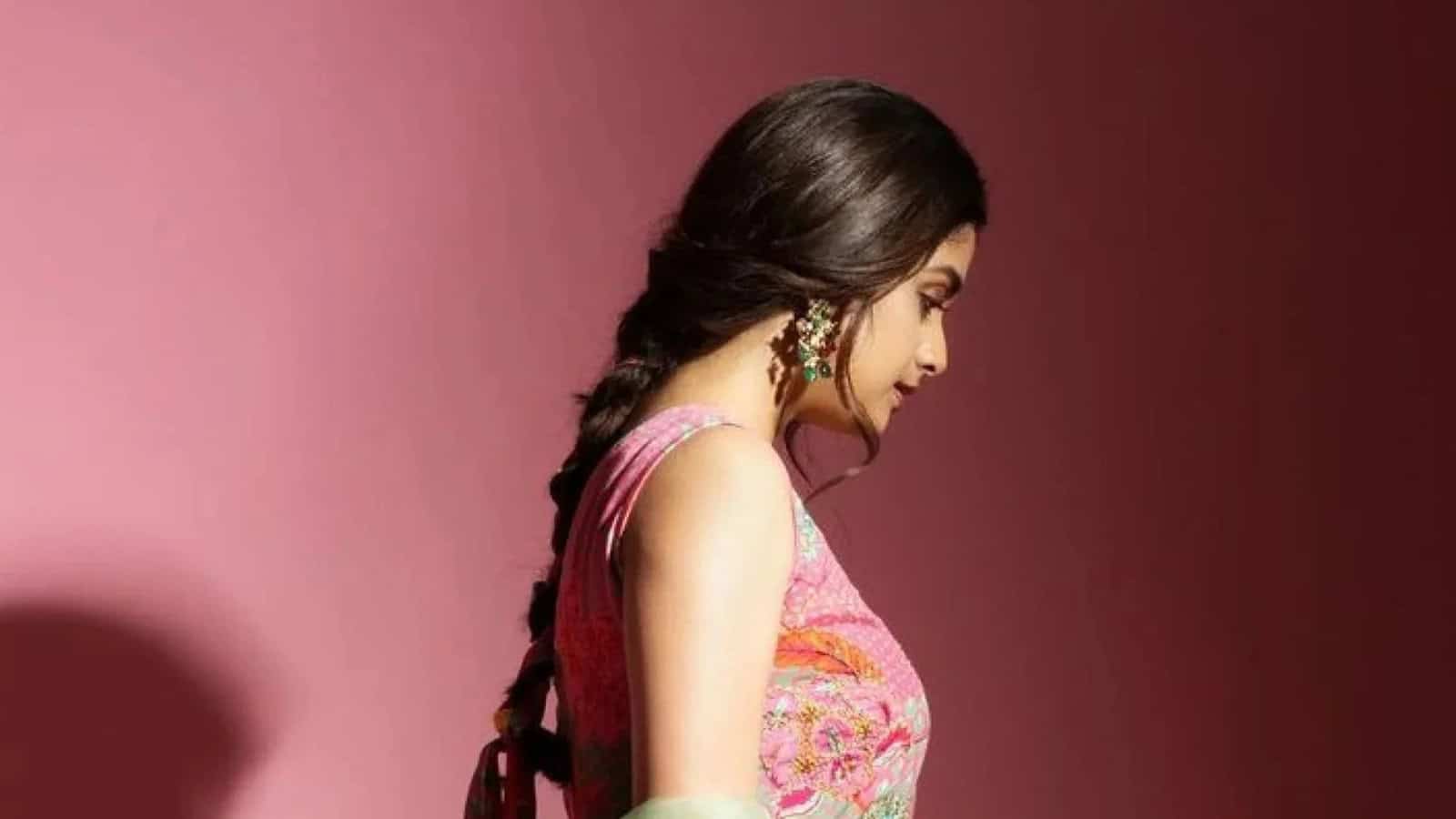 Woman in traditional dress with braided hair, looking down against pink backdrop.
