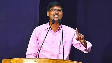 Man speaking at a conference, wearing pink shirt, gesturing with his finger, standing behind a podium wit.