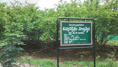 Signboard in Telangana urging protection of seed farm lands, surrounded by lush green trees.