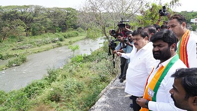 Politicians and media at riverbank during environmental cleanup activity.