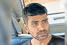 Man with short black hair and beard sitting in a vehicle, looking serious.