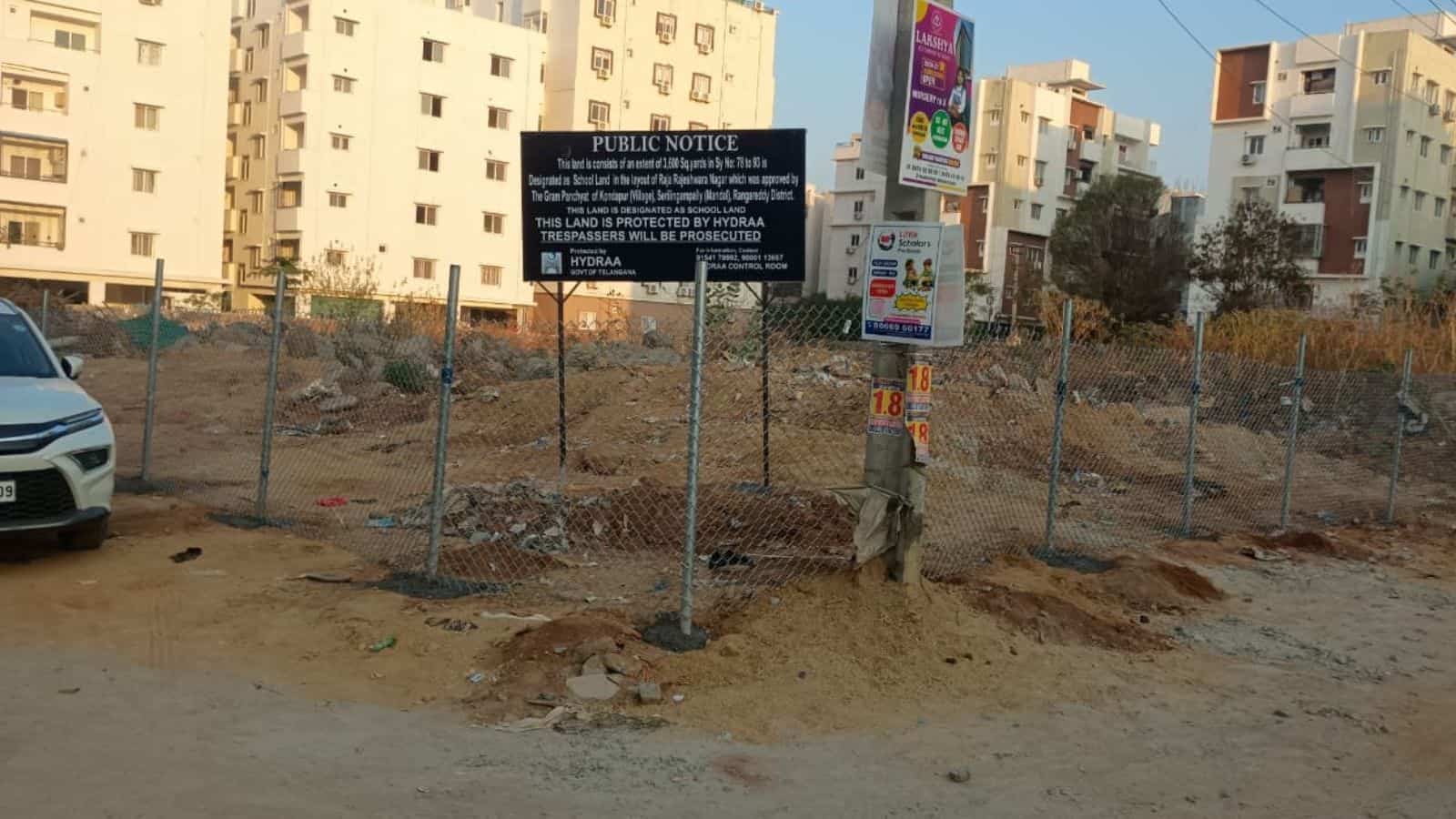 Fenced construction area in Hyderabad with urban buildings and a public notice sign.