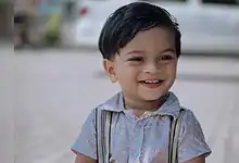 Smiling young boy with dark hair wearing a light shirt and suspenders outdoors.