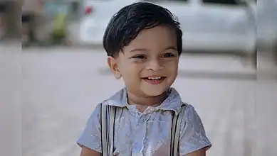 Smiling young boy with dark hair wearing a light shirt and suspenders outdoors.