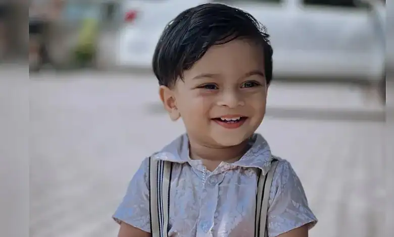 Smiling young boy with dark hair wearing a light shirt and suspenders outdoors.