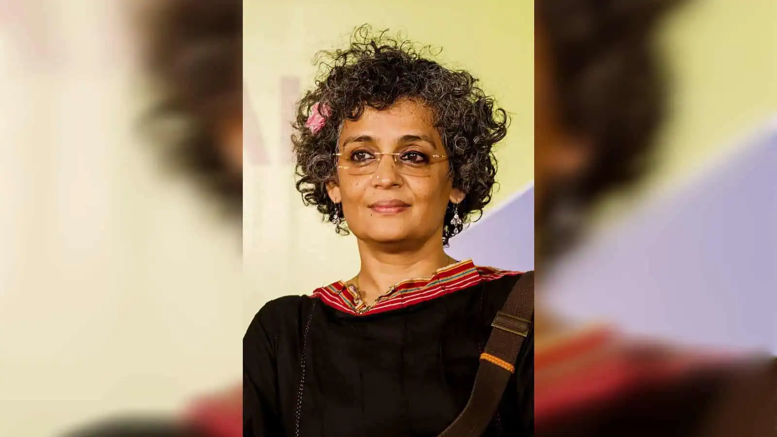 Arundhati Roy at a public event, wearing glasses and a black outfit with a red-bordered neckline.