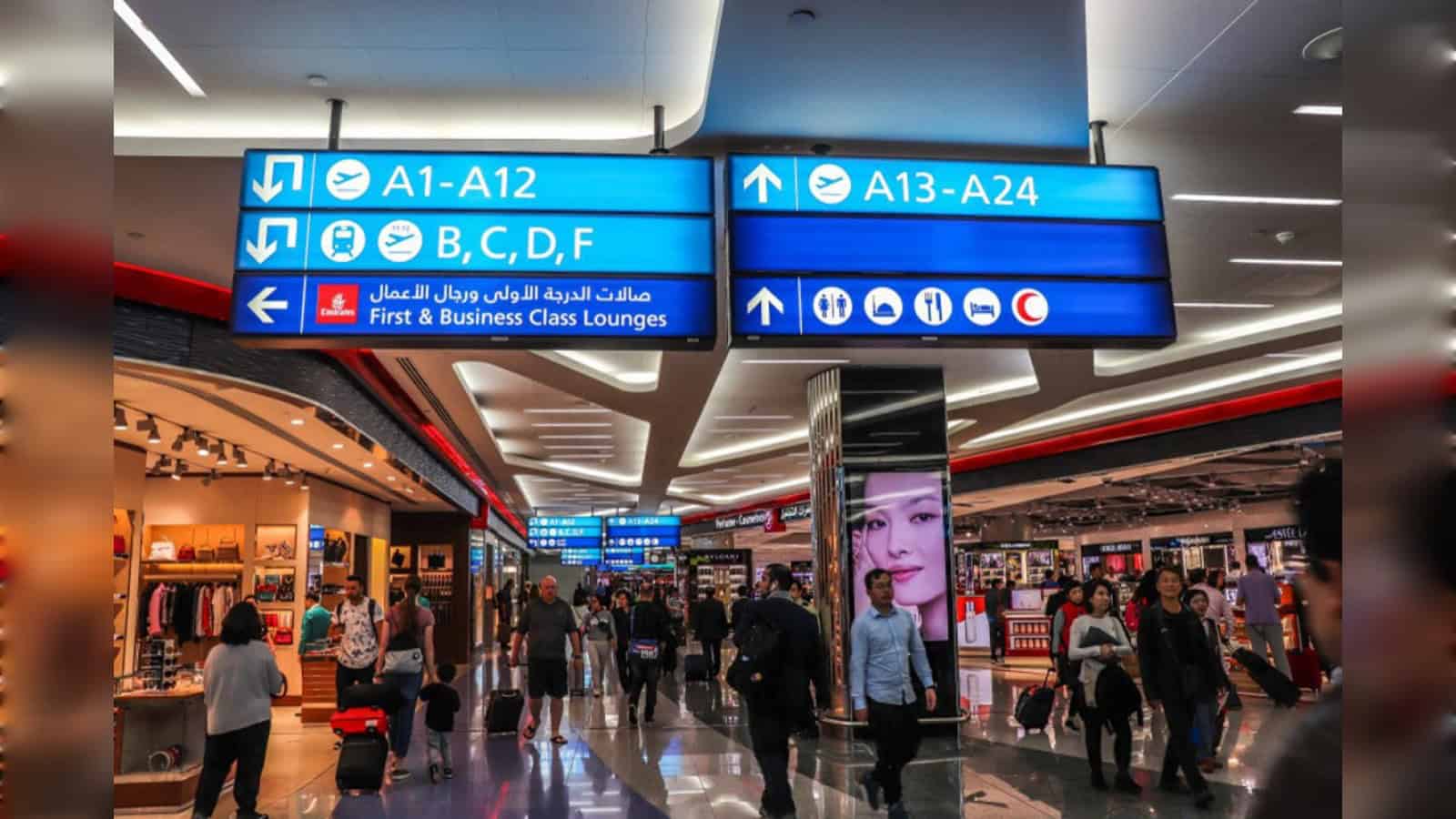 Passengers walk through a departure terminal at Dubai International Airport with gate signs overhead.