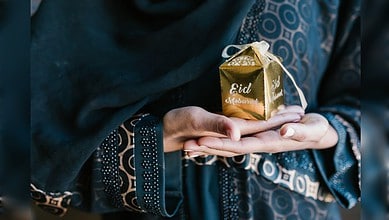 Person holding a gold gift box with "Eid Mubarak" in traditional attire, celebrating Eid.