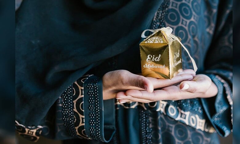 Person holding a gold gift box with "Eid Mubarak" in traditional attire, celebrating Eid.
