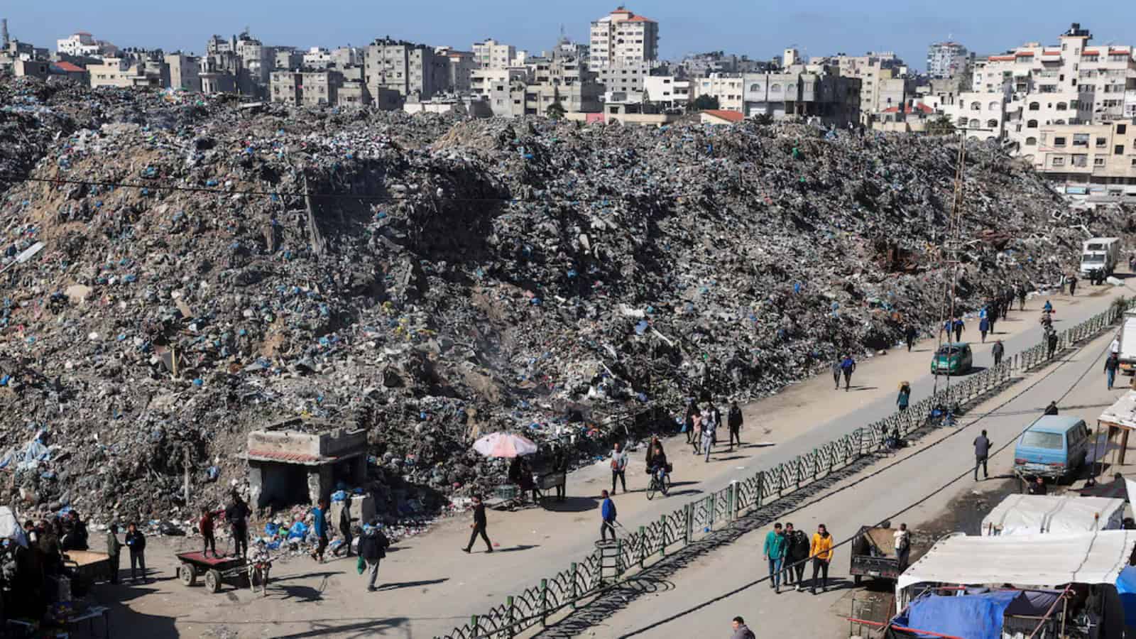 Large garbage dump in Gaza with residents walking along the street.