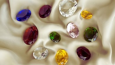 Image shows a collection of faceted gemstones in various colors including blue, purple, red, clear, yellow, pink, and green arranged on champagne-colored silk fabric.