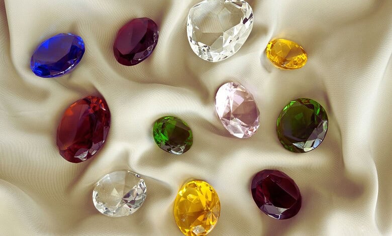 Image shows a collection of faceted gemstones in various colors including blue, purple, red, clear, yellow, pink, and green arranged on champagne-colored silk fabric.