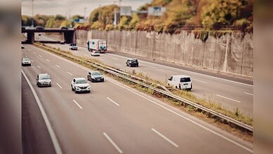Image shows Multi-lane highway with light traffic including cars, trucks, and vans traveling in both directions, bordered by concrete retaining walls and vegetation with a tilt-shift photography effect.