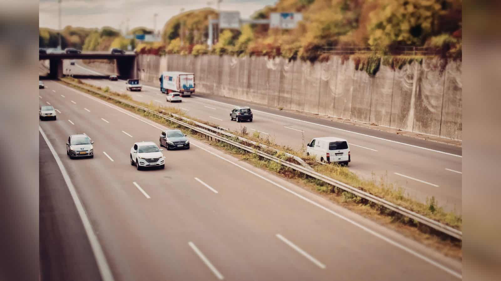 Image shows Multi-lane highway with light traffic including cars, trucks, and vans traveling in both directions, bordered by concrete retaining walls and vegetation with a tilt-shift photography effect.