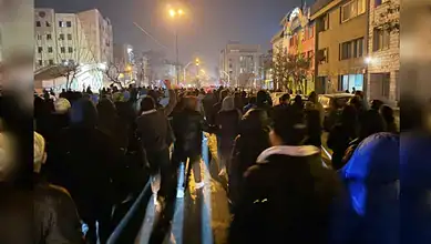Large crowd gathered on a city street at night, illuminated by streetlights and building lights.