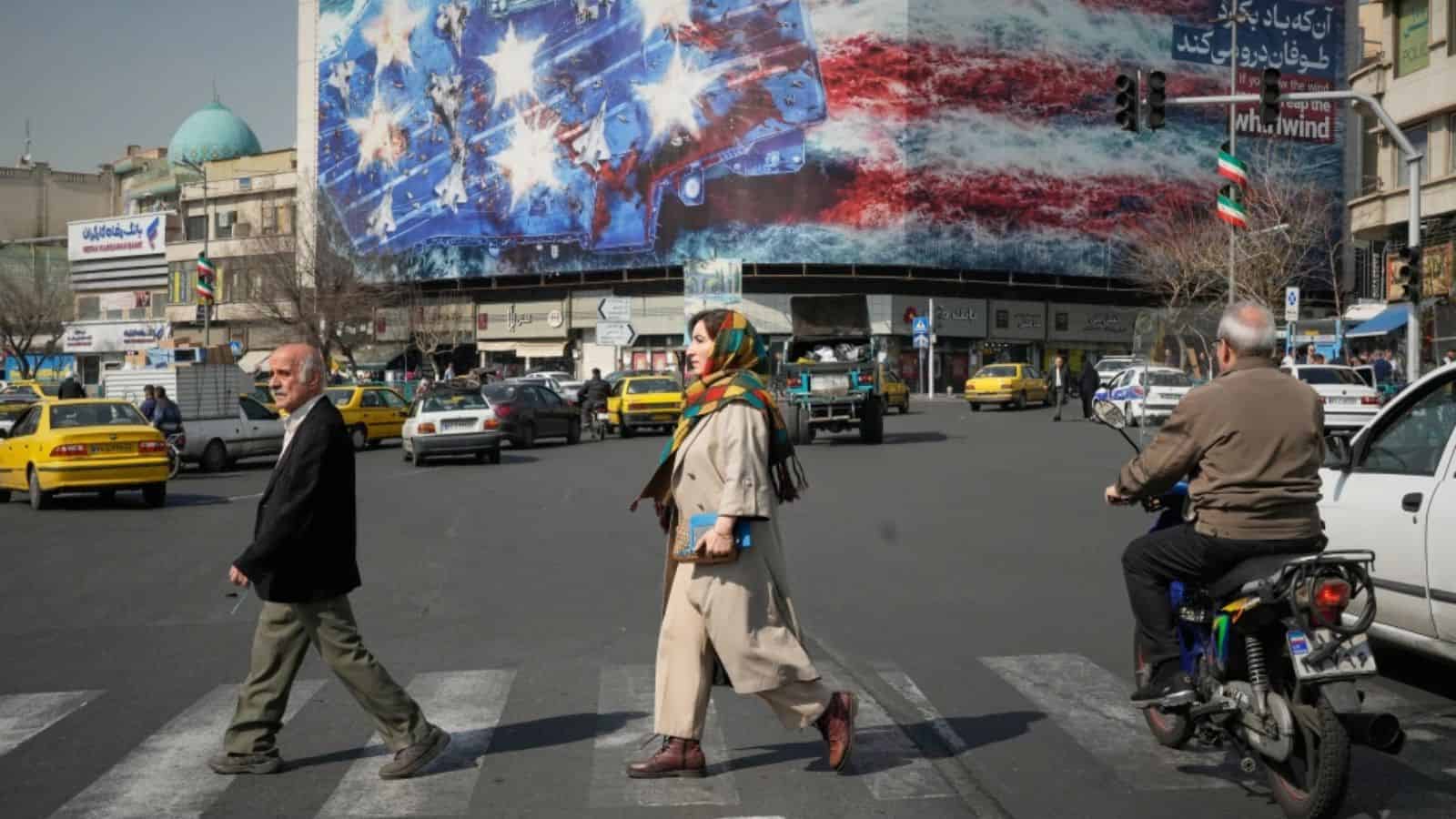 People crossing a busy street in Iran with a large American flag and protest banners in the background.