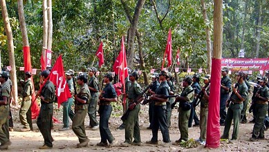 Image shows Armed Maoist insurgents in military-style uniforms carrying red flags march through a forested area during a gathering.