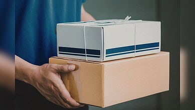 Person holding two parcels, one in a cardboard box and one in a white box, related to rail parcel service.