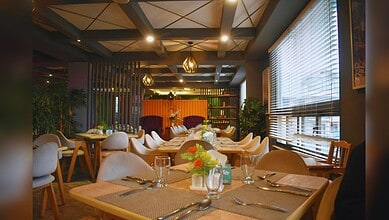 Modern restaurant interior featuring warm wood tables with white chairs, geometric coffered ceiling, and natural light from large windows