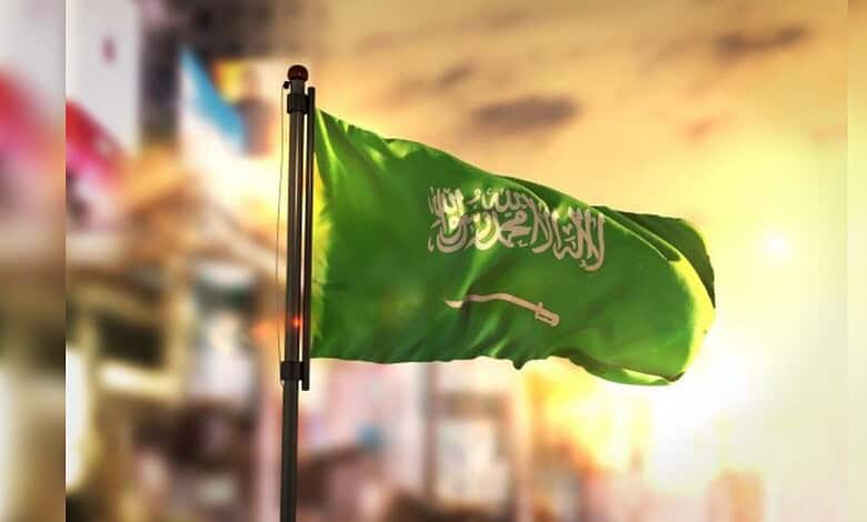 Saudi Arabian flag waving on a pole against a city skyline at sunset.