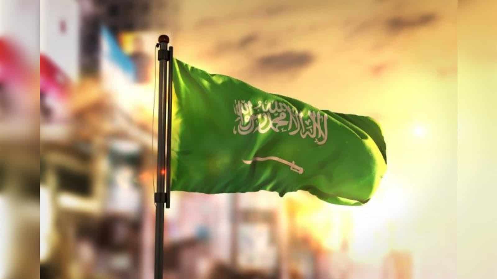 Saudi Arabian flag waving on a pole against a city skyline at sunset.