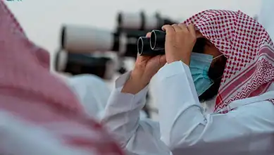 Saudi moon sighting committee member uses binoculars to observe the sky while wearing traditional attire and a face mask.
