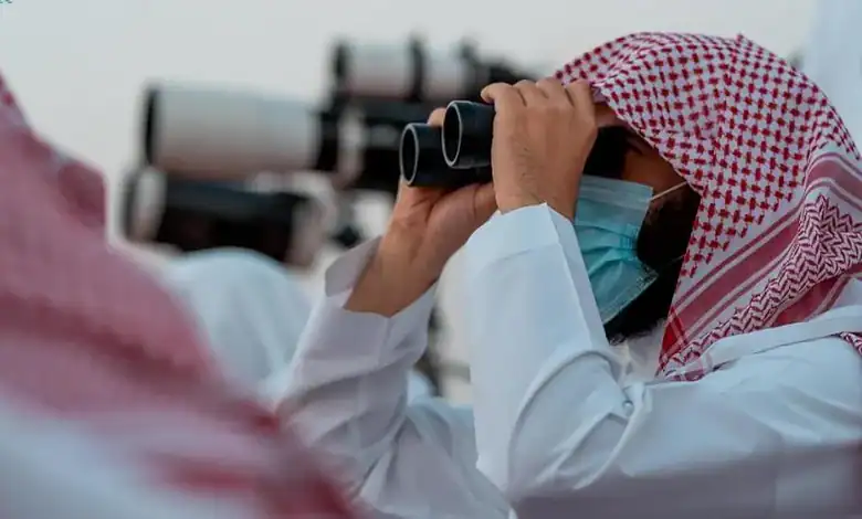 Saudi moon sighting committee member uses binoculars to observe the sky while wearing traditional attire and a face mask.