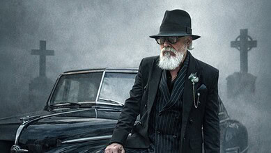 Elderly man with white beard, sunglasses, and black hat standing beside vintage car in foggy graveyard.