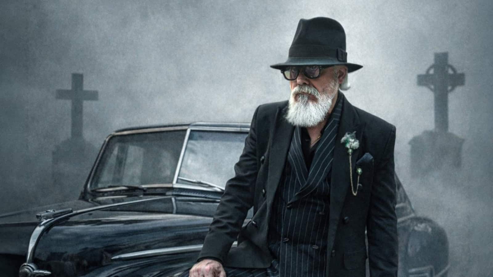 Elderly man with white beard, sunglasses, and black hat standing beside vintage car in foggy graveyard.
