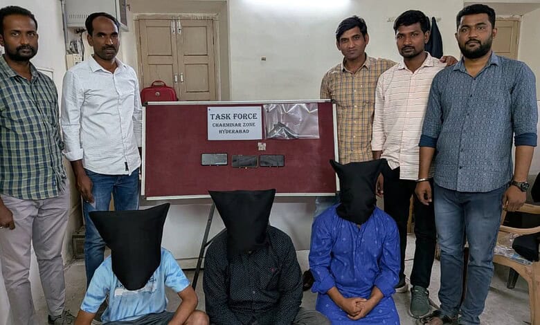 Group of five men standing with two suspects seated with hoods over their heads in an office setting.