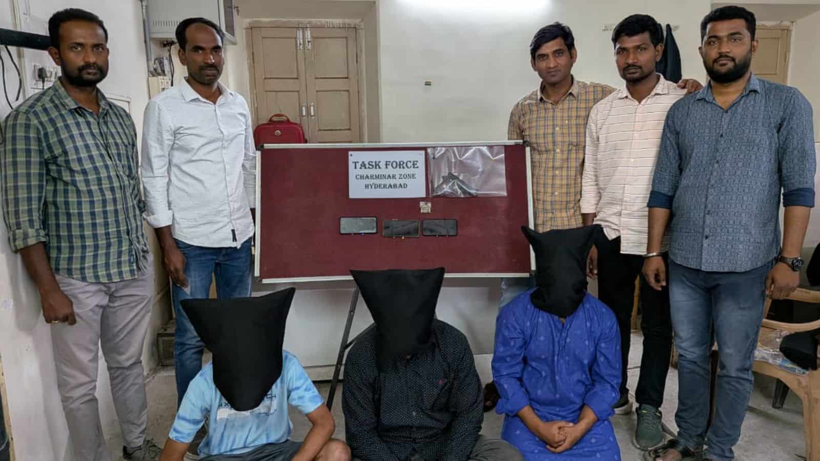 Group of five men standing with two suspects seated with hoods over their heads in an office setting.