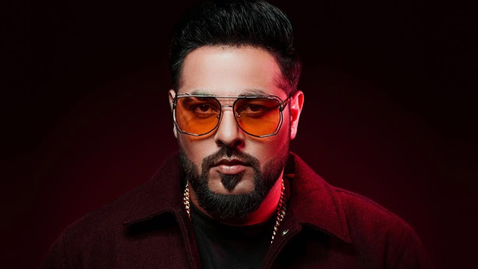 Portrait of Badshah with intense expression, dark background, and stylish sunglasses.