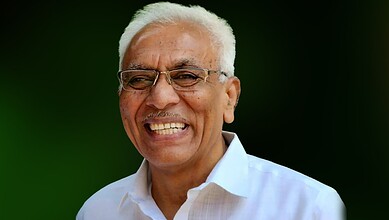 Smiling Indian politician with white hair and glasses, wearing a white shirt, against a green background.