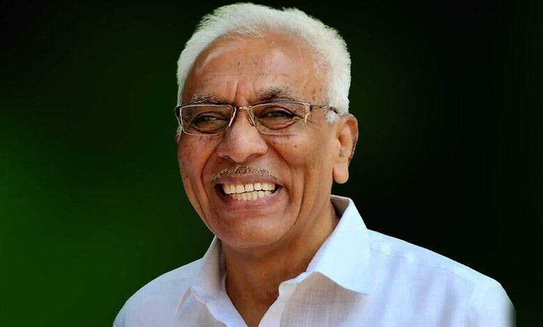 Smiling Indian politician with white hair and glasses, wearing a white shirt, against a green background.