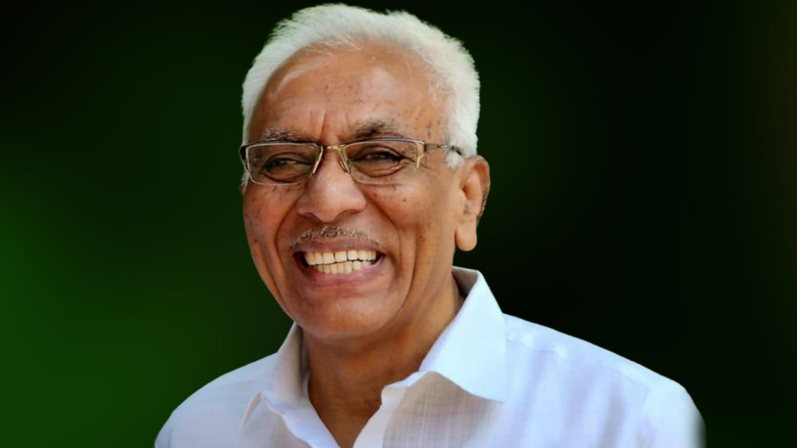 Smiling Indian politician with white hair and glasses, wearing a white shirt, against a green background.