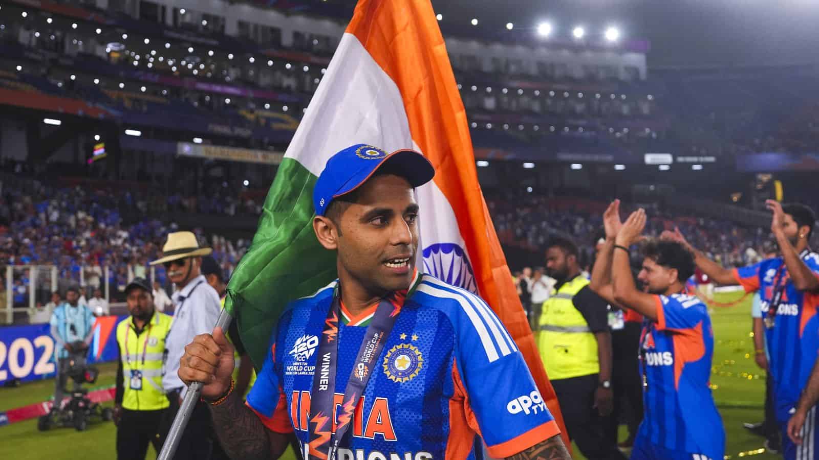 Suryakumar Yadav holding the Indian flag after a cricket match victory, surrounded by teammates and fans.