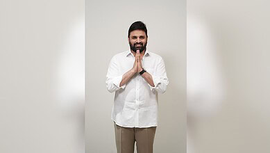 An image of a smiling man with hands folded in a gesture of greeting or respect, standing against a plain.