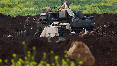 Military tank and soldiers in a field, with green grass and soil, possibly during a training or conflict.