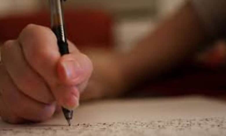 Student's hand holding a pen and writing on exam paper during class 12 board exams.