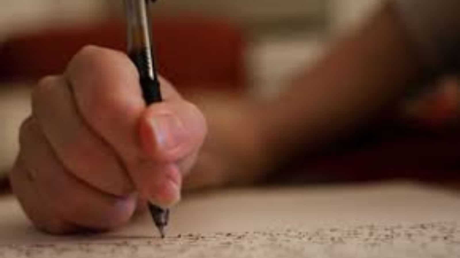 Student's hand holding a pen and writing on exam paper during class 12 board exams.