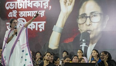 Mamata Banerjee addressing a rally, gesturing with her hand, with a large poster of herself behind her.