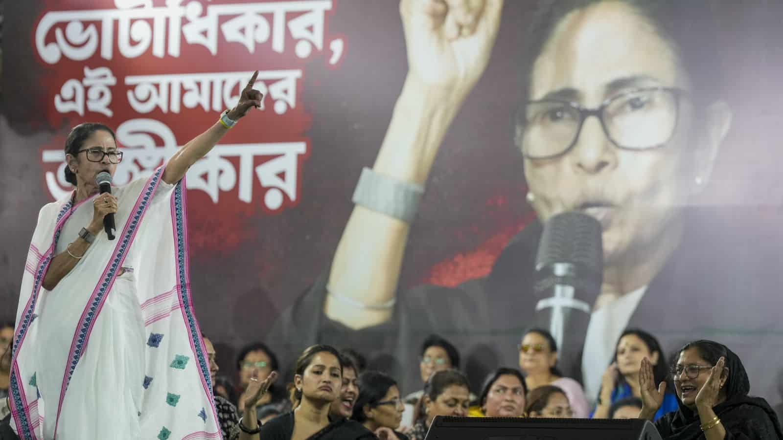 Mamata Banerjee addressing a rally, gesturing with her hand, with a large poster of herself behind her.