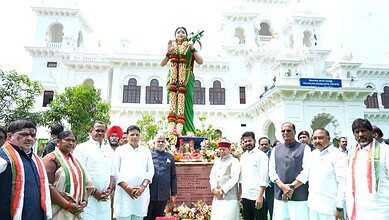 Telangana CM addressing a public gathering with a statue of a woman leader in the background.