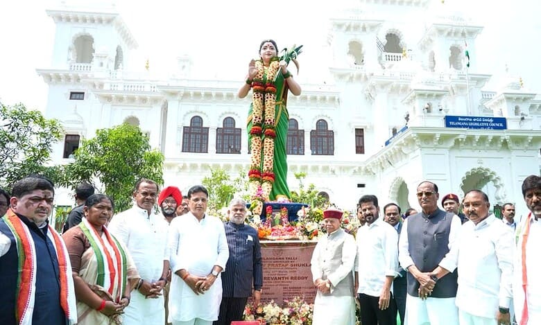 Telangana CM addressing a public gathering with a statue of a woman leader in the background.