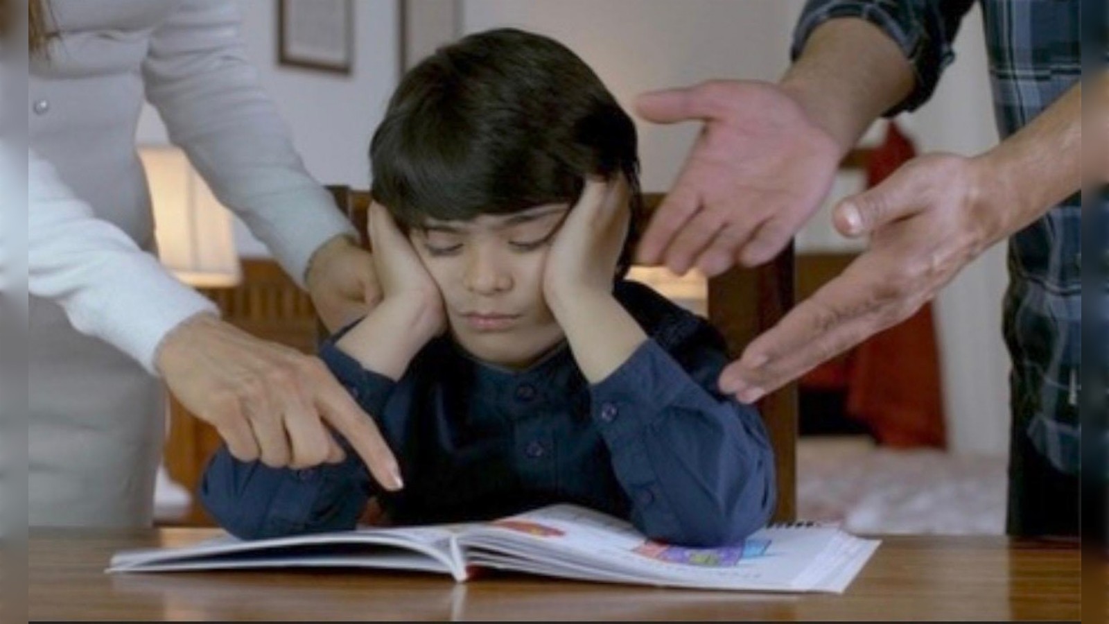 Representative image of a child covering ears while adults point at a book, appearing stressed during study.