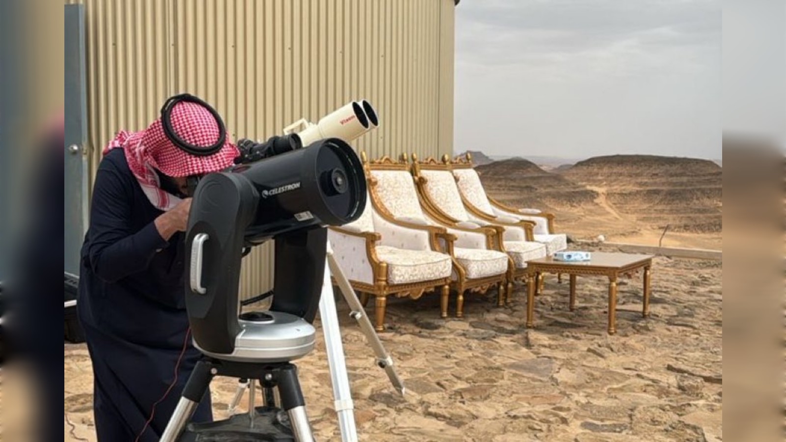 Man uses a telescope to sight the crescent moon in a desert setting.