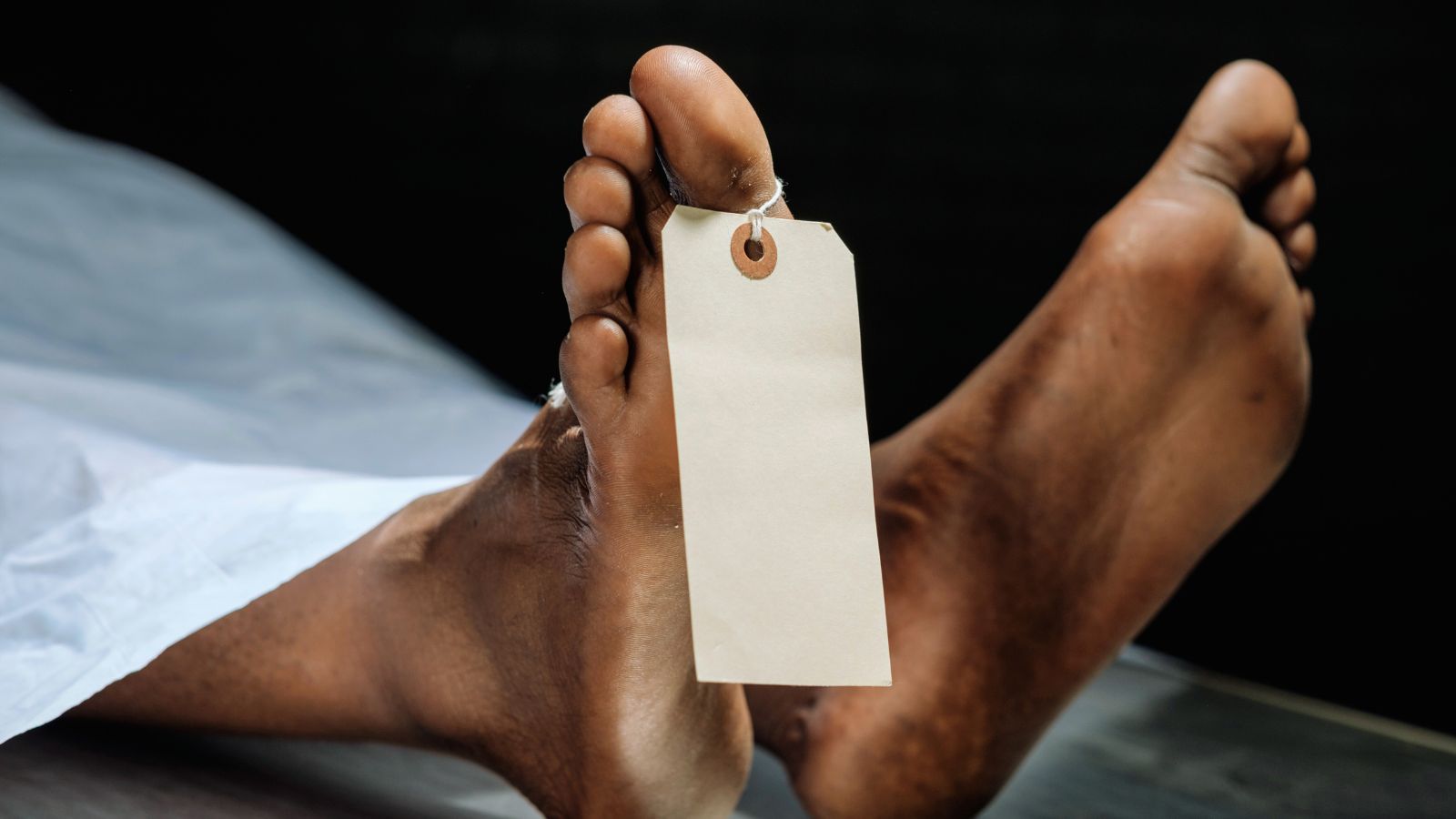 Image shows Close-up of a deceased person's feet with a blank identification tag tied to the toe in a morgue setting.