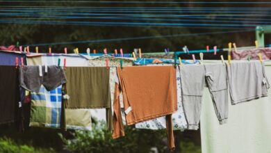 Colorful clothes and linens hanging on parallel blue clotheslines outdoors, secured with multicolored wooden and plastic clothespins, with green foliage in the background.