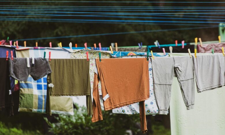 Colorful clothes and linens hanging on parallel blue clotheslines outdoors, secured with multicolored wooden and plastic clothespins, with green foliage in the background.