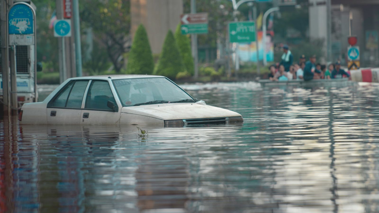 Image shows A white car nearly submerged in floodwater on a city street as onlookers watch from higher ground nearby.